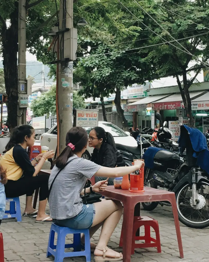 Friends on low plastic stools at a pavement cafe on a leafy Da Nang street, motorbikes parked behind them