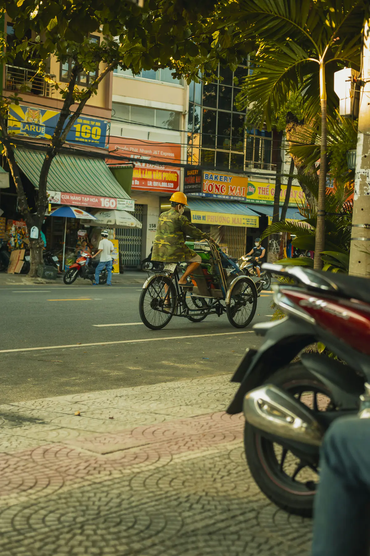 A cyclo driver in a yellow helmet pedals past bakery and shop signs on a Da Nang street in late afternoon light